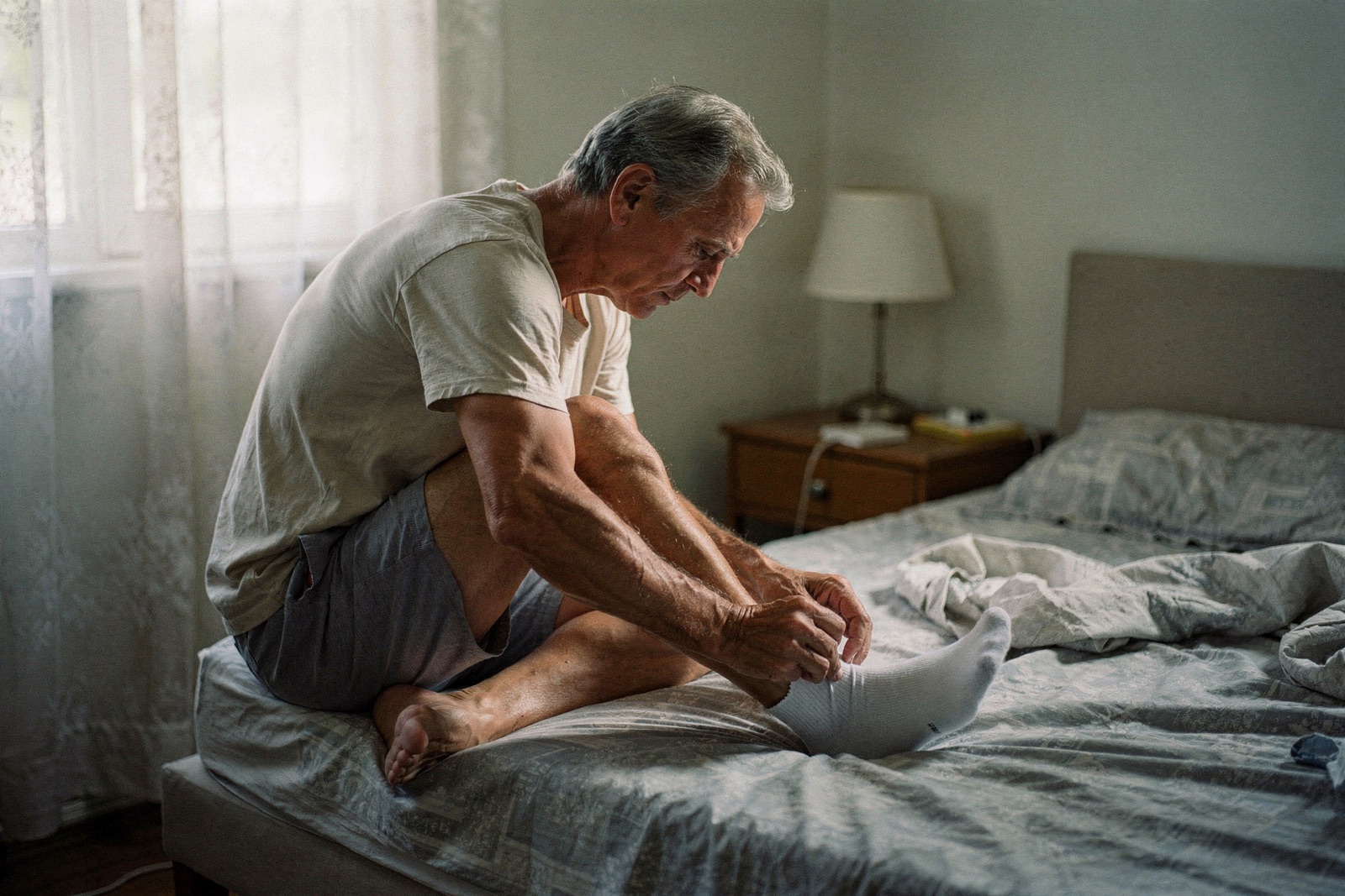 A man in his sixties sits on the edge of his bed in morning light, pulling on a clean cotton sock.