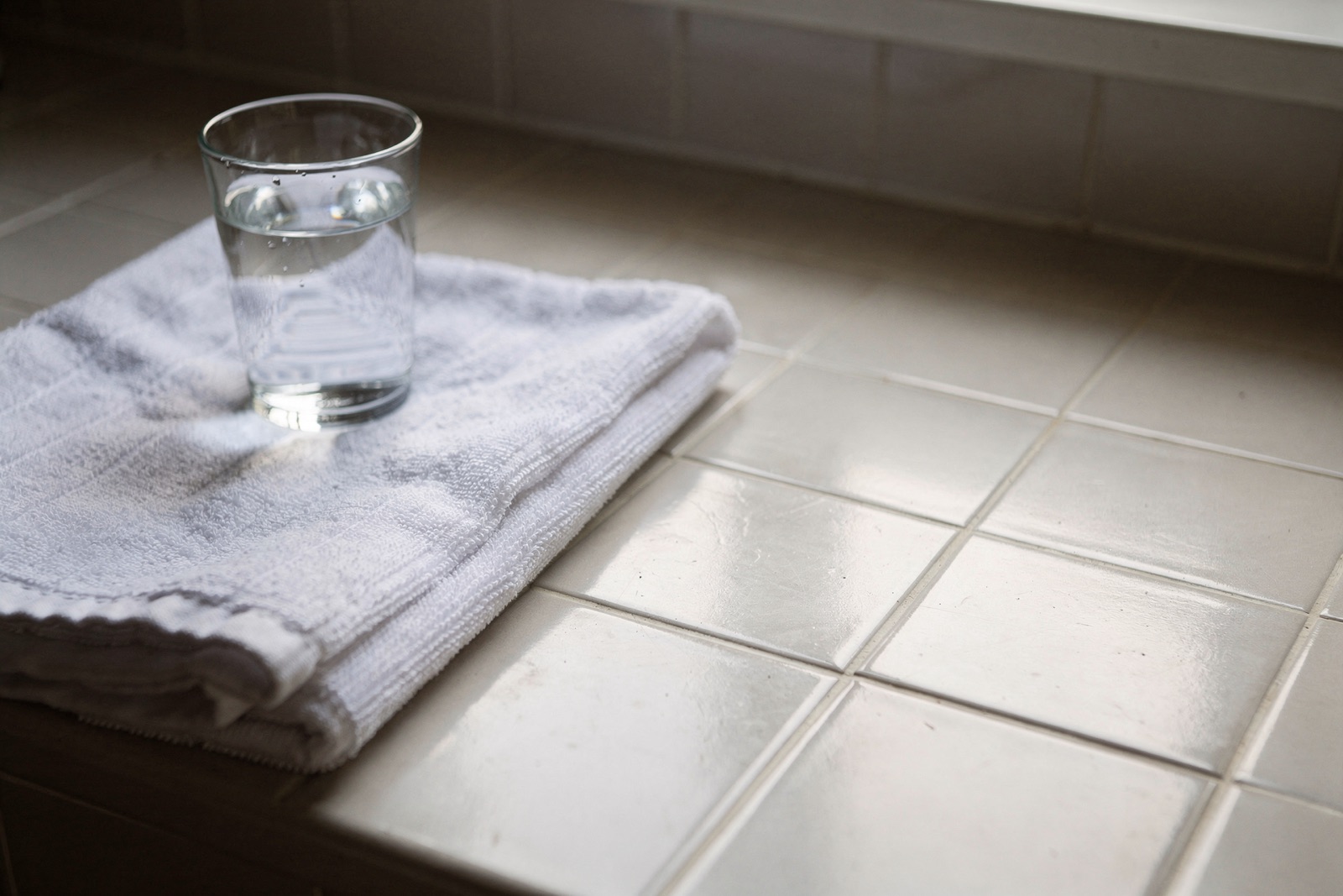 A tiled bathroom counter in morning daylight with a folded white hand towel and a small glass of water.